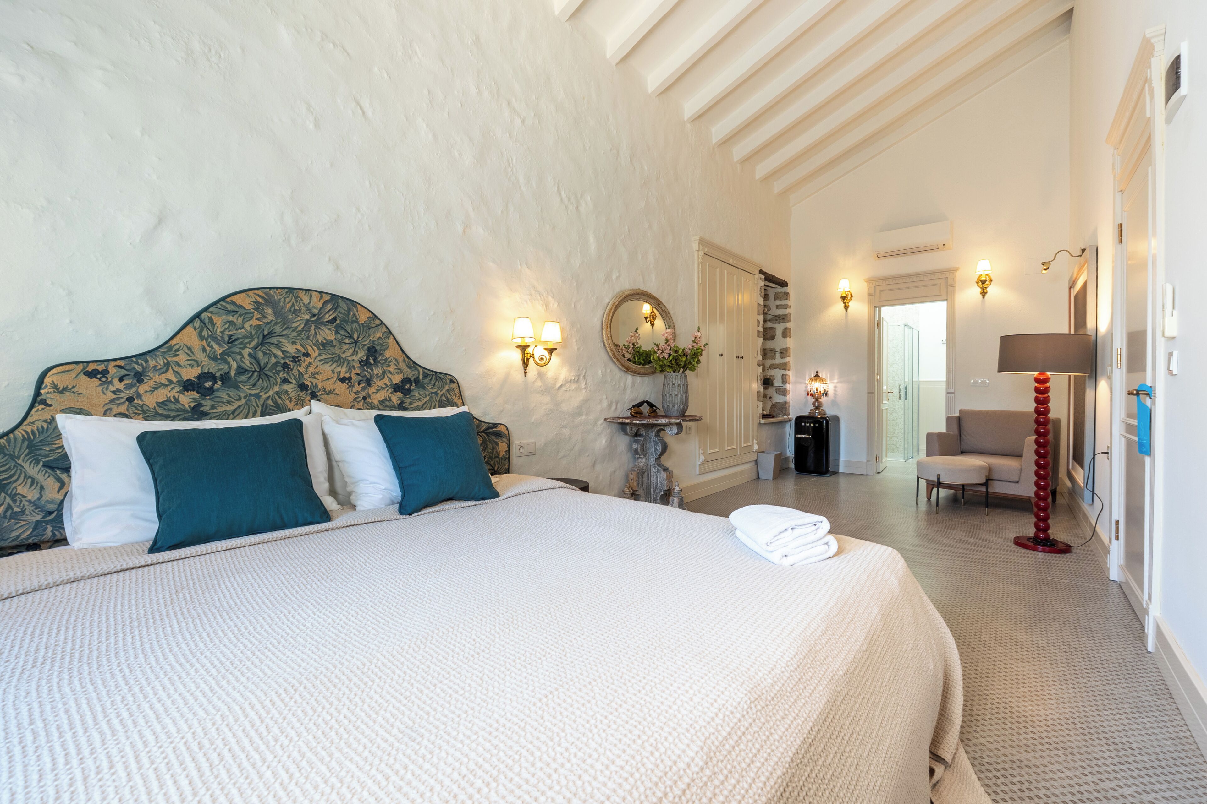 A bedroom with a large bed, a headboard with a floral pattern, two wall-mounted lamps, a round mirror, and a small table with a vase.