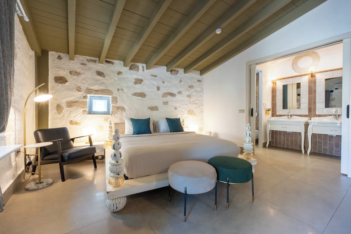 Ein Schlafzimmer mit Steinsandsteinwand, einem Bett, einem Sessel, einem runden Hocker, einem Schminktisch mit Spiegel und einem durchgegangenen Tür erkennbaren Badezimmer.