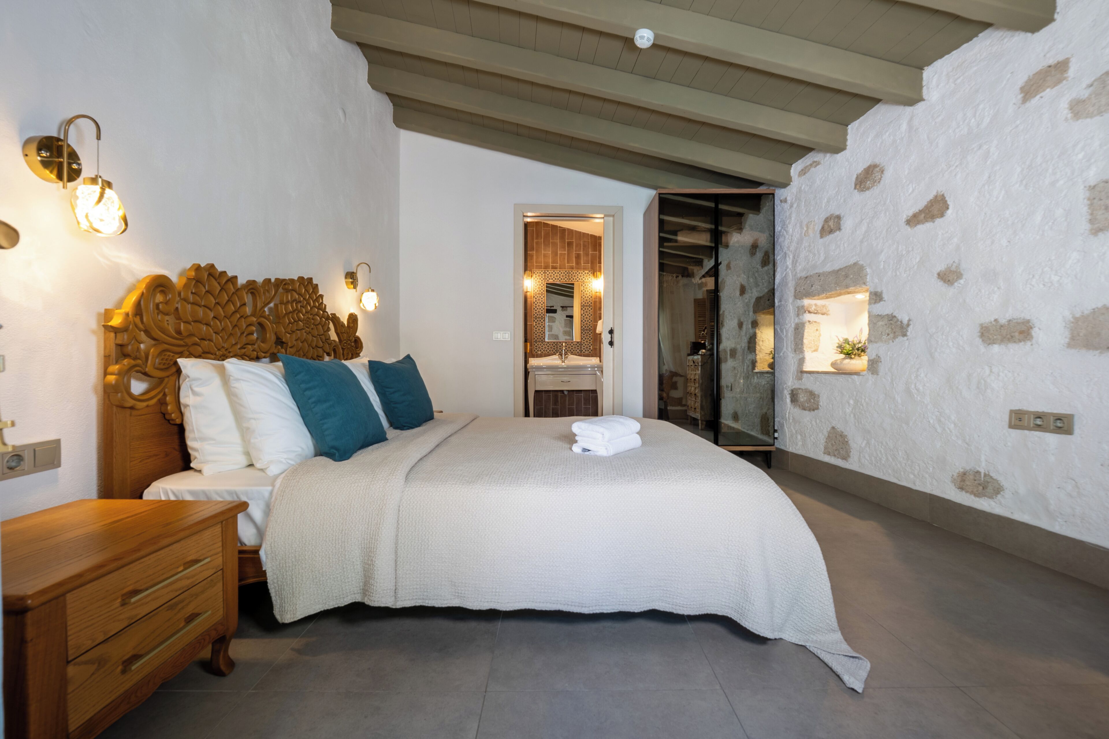 A bedroom with a wooden headboard, a large bed, a nightstand, and a stone wall.