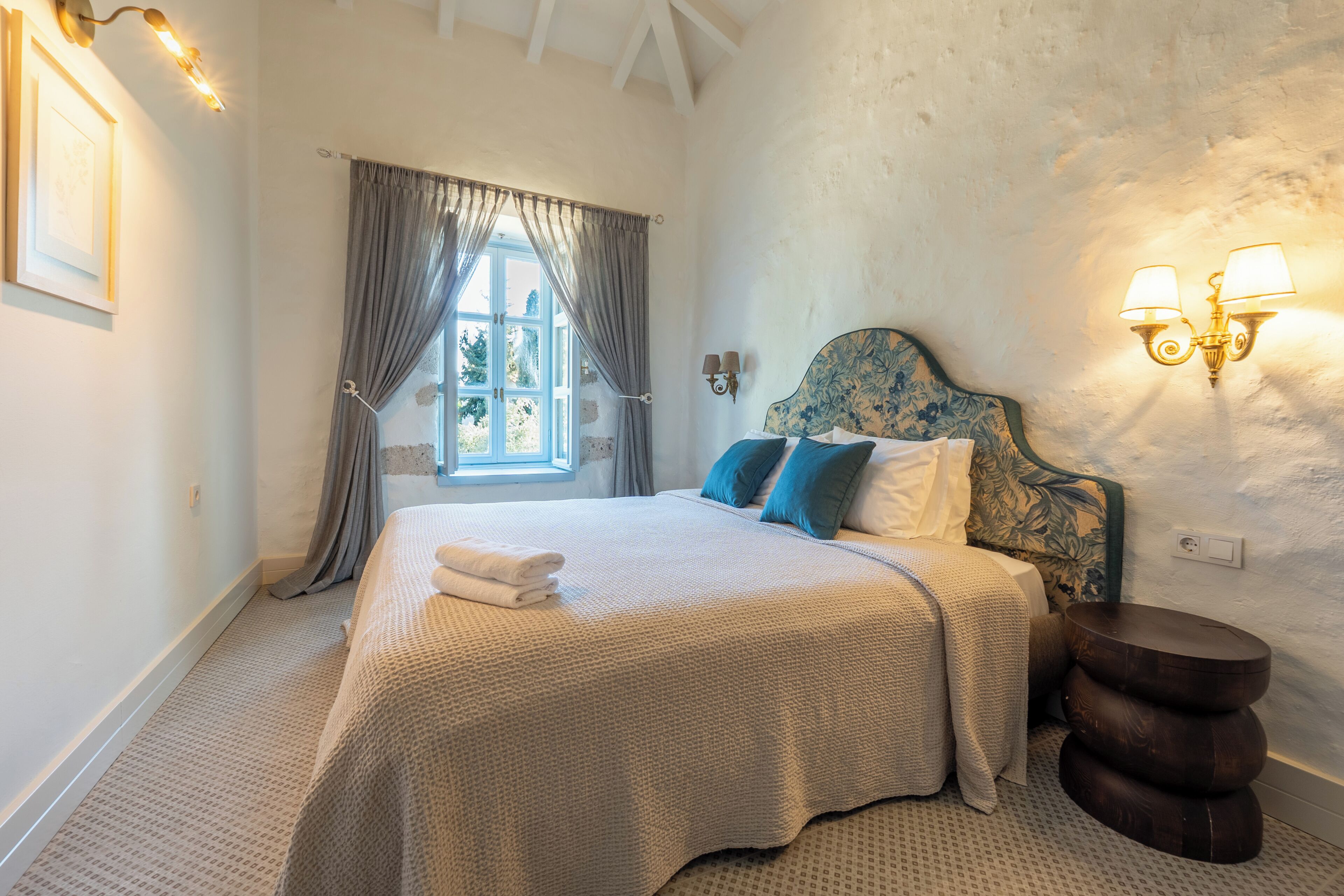 Ein Schlafzimmer mit Bett, Vorhängen, einem Fenster, Wandleuchten und einem runden Nachttisch aus Holz.