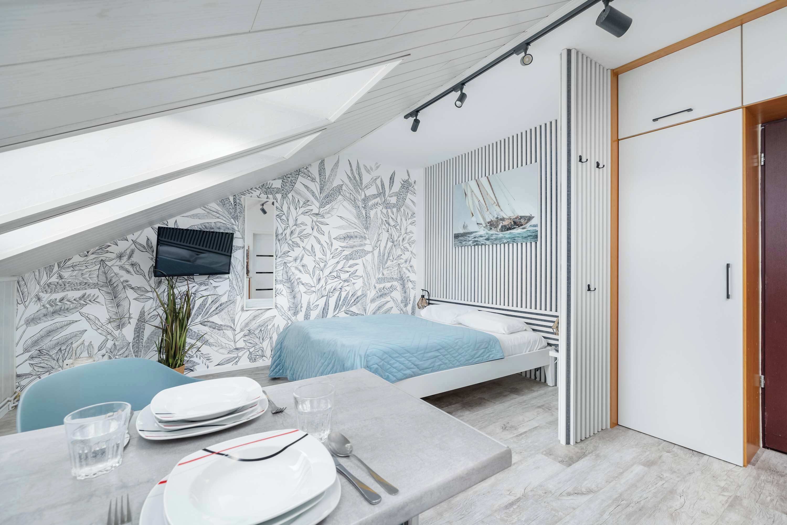 Ein modernes Schlafzimmer mit schräg verlaufender Decke, einem Bett mit gestreifter Kopfteil, einem an der Wand montierten Fernseher und einem großen Fenster.