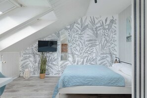 Ein Schlafzimmer mit schrägen Wänden, einem Bett mit blauer Decke, einem an der Wand befestigten Fernseher und einem großen Fenster.