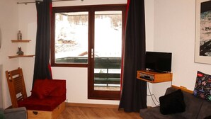 Ein gemütlicher Wohnraum mit Holzboden, einem roten Sessel, einem grauen Sofa, einem Fernseher auf einer Holzablage und einem Fenster mit Vorhängen.