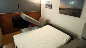 A compact room with a bed, a desk, and a chair. There is a wall-mounted picture and a lamp.