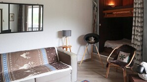 Ein gemütliches Wohnzimmer mit einer Couch, einem Sessel, einem kleinen Tisch und einer Lampe. Im Hintergrund ist ein Bett mit einem Holz-Kopfstück zu sehen.