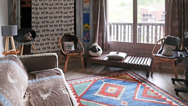 Ein gemütliches Wohnzimmer mit einer Couch, Sesseln, einem Teppich und einem Fenster mit Blick auf eine Holzterrasse und Gebäude.