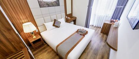 A hotel room with a large bed, wooden headboard, a bedside table with a lamp, a mirror, and a window with curtains.