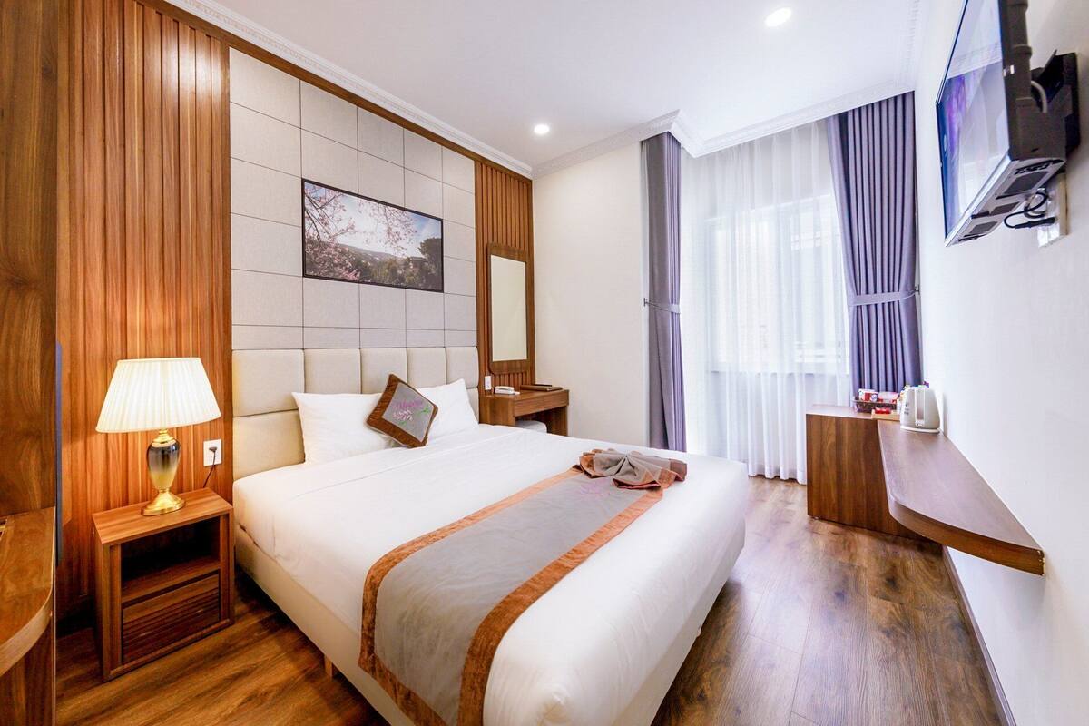 Une chambre d’hôtel avec un grand lit, une table de chevet avec une lampe, un téléviseur fixé au mur et une fenêtre avec des rideaux.
