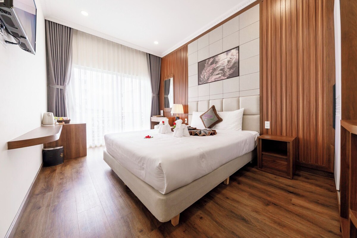 A hotel room with a large bed, wooden paneling, a flat-screen TV, a bedside table, and a window with curtains.