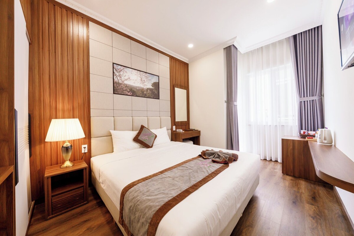 A hotel room with a large bed, a bedside table with a lamp, a wooden headboard, a painting on the wall, a desk with a chair, and a window with curtains.