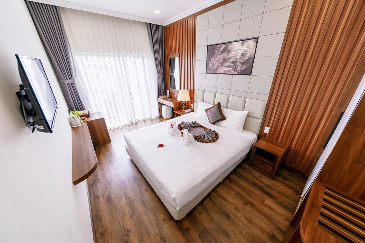 A hotel room with a bed, a TV mounted on the wall, a bedside table with a lamp, and a large window with curtains.