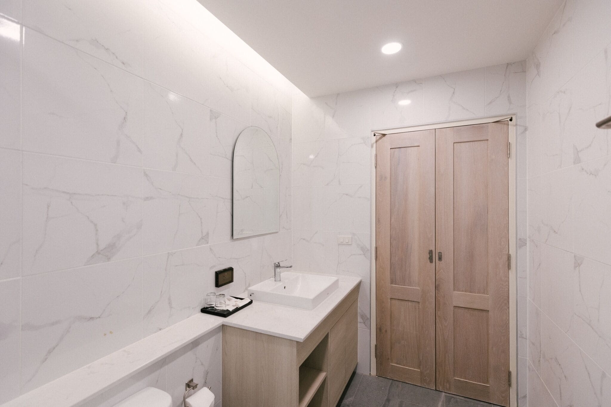 A modern bathroom with white marble walls, a wooden vanity, a large mirror, and a wooden door.