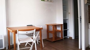 Ein kleiner Raum mit einem Holztisch, einem weißen Stuhl, einem an der Wand befestigten Fernseher und einer teilweise geöffneten Tür zu einem Schrank.