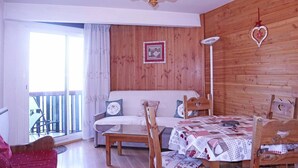 A cozy room with a wooden wall, a chandelier, a sofa with cushions, a dining table with a quilt, and a window with a view.