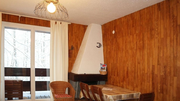 Una habitación acogedora con paredes de madera, una mesa de comedor con sillas, un sillón de mimbre, una chimenea y un ventanal con cortinas.