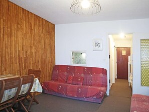 A room with a wooden wall, a red sofa, a dining table with chairs, a small window, and a door leading to another room.