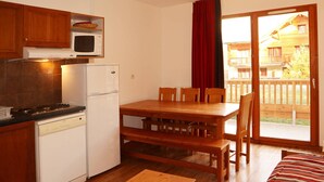A compact kitchen with a stove, microwave, and refrigerator, a wooden dining table with chairs, and a sliding glass door leading to a balcony. - Spacious 3-Room Apartment for 8 with Heated Pool & Terrace in Les Orres 1800 (Les Orres)