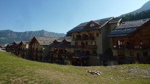 A row of wooden chalet-style buildings with balconies, set against a mountainous backdrop. - Spacious 3-Room Apartment for 8 with Heated Pool & Terrace in Les Orres 1800 (Les Orres)