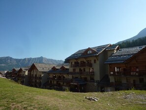 A row of wooden chalet-style buildings with balconies, set against a mountainous backdrop. - Spacious 3-Room Apartment for 8 with Heated Pool & Terrace in Les Orres 1800 (Les Orres)