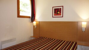 A single bed with a striped bedspread, a wooden headboard, two bedside lamps, a window with a curtain, and a framed picture on the wall. - Spacious 3-Room Apartment for 8 with Heated Pool & Terrace in Les Orres 1800 (Les Orres)