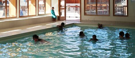 An indoor swimming pool with clear glass walls, wooden deck outside, and tiled floor.