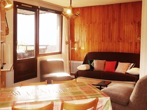 A cozy room with wooden paneling, a sofa with cushions, a dining table with chairs, and a window with curtains.