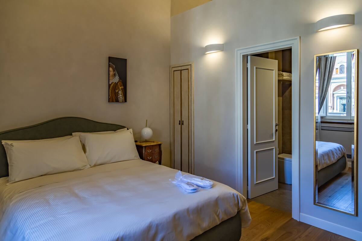 Habitación de hotel con cama, mesita de noche, un cuadro, armario y un espejo que refleja la habitación.
