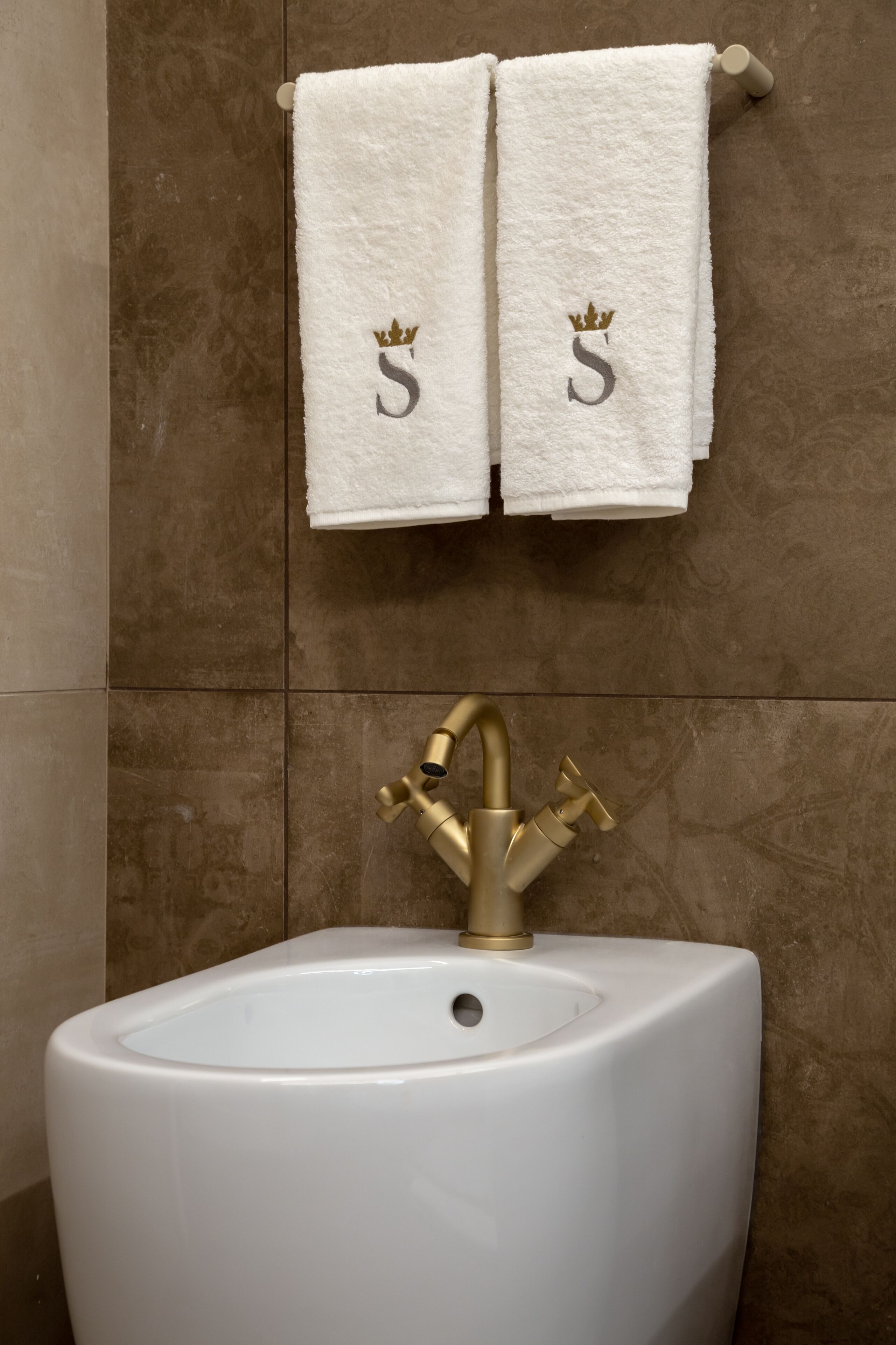 Une salle de bain avec deux serviettes blanches accrochées au mur, chacune ornée d’une couronne dorée et de la lettre « S ». En dessous des serviettes, on trouve un lavabo blanc avec un robinet doré.