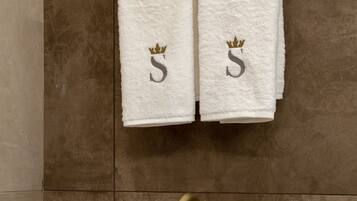 Une salle de bain avec deux serviettes blanches accrochées au mur, chacune ornée d’une couronne dorée et de la lettre « S ». En dessous des serviettes, on trouve un lavabo blanc avec un robinet doré.