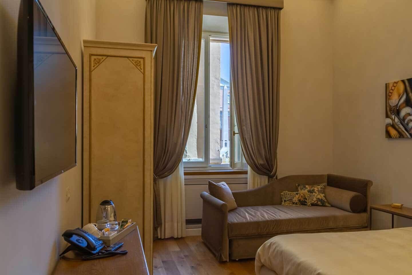 Habitación de hotel con cama, sofá, escritorio con teléfono y vaso, y ventana con cortinas.