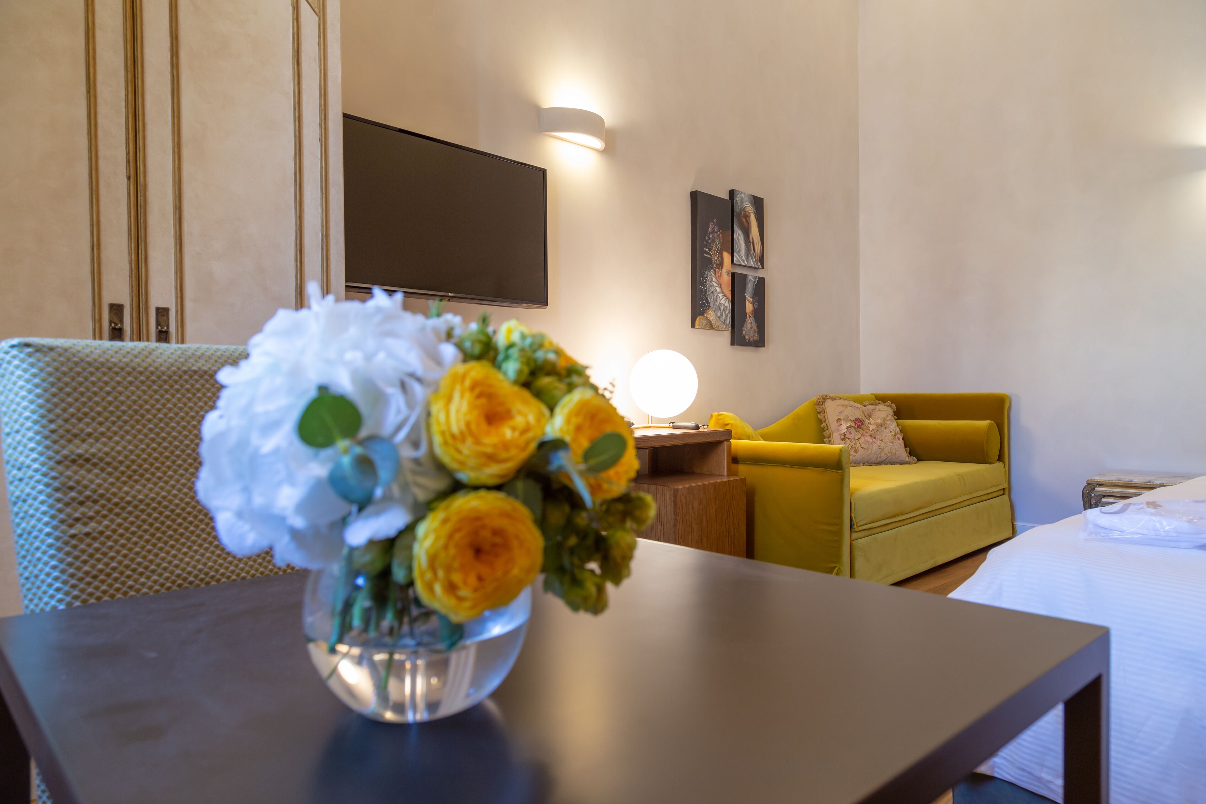 A hotel room with a bed, a yellow armchair, a small table with a vase of flowers, and a flat-screen TV mounted on the wall.
