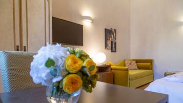A hotel room with a bed, a yellow armchair, a small table with a vase of flowers, and a flat-screen TV mounted on the wall.