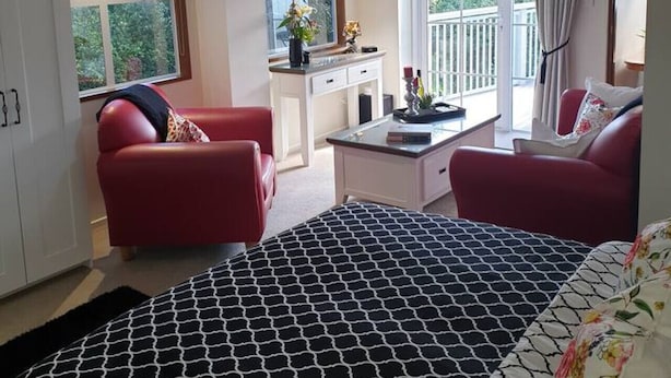 A bedroom with a large bed, two red sofas, a small desk, and a balcony with a view of greenery. - Tui2 - Studio (Macandrew Bay)