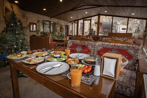 A dining area with a wooden table set for breakfast, a Christmas tree, and a view of the mountains through large windows. - Zeus Cave Suites (Nevsehir)