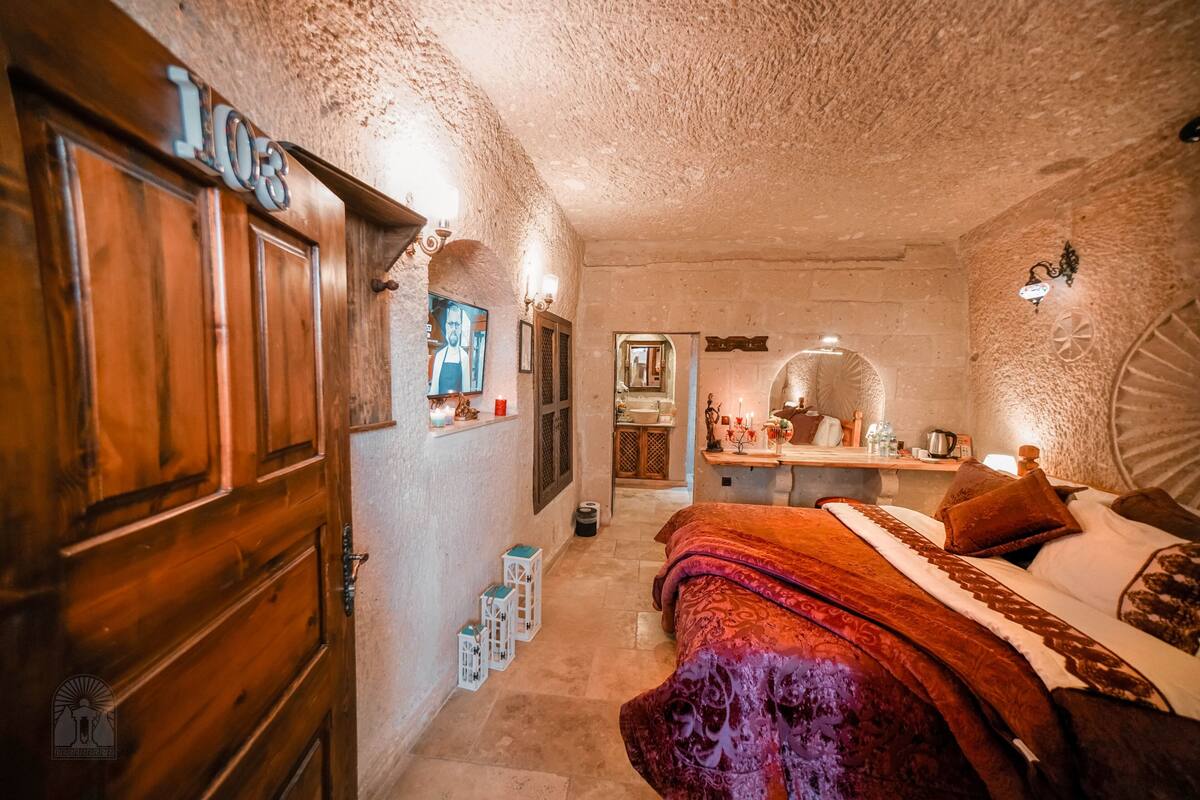 A cozy, rustic bedroom with a wooden door labeled "103", a bed with a red and purple blanket, and a small desk with a mirror and various items.