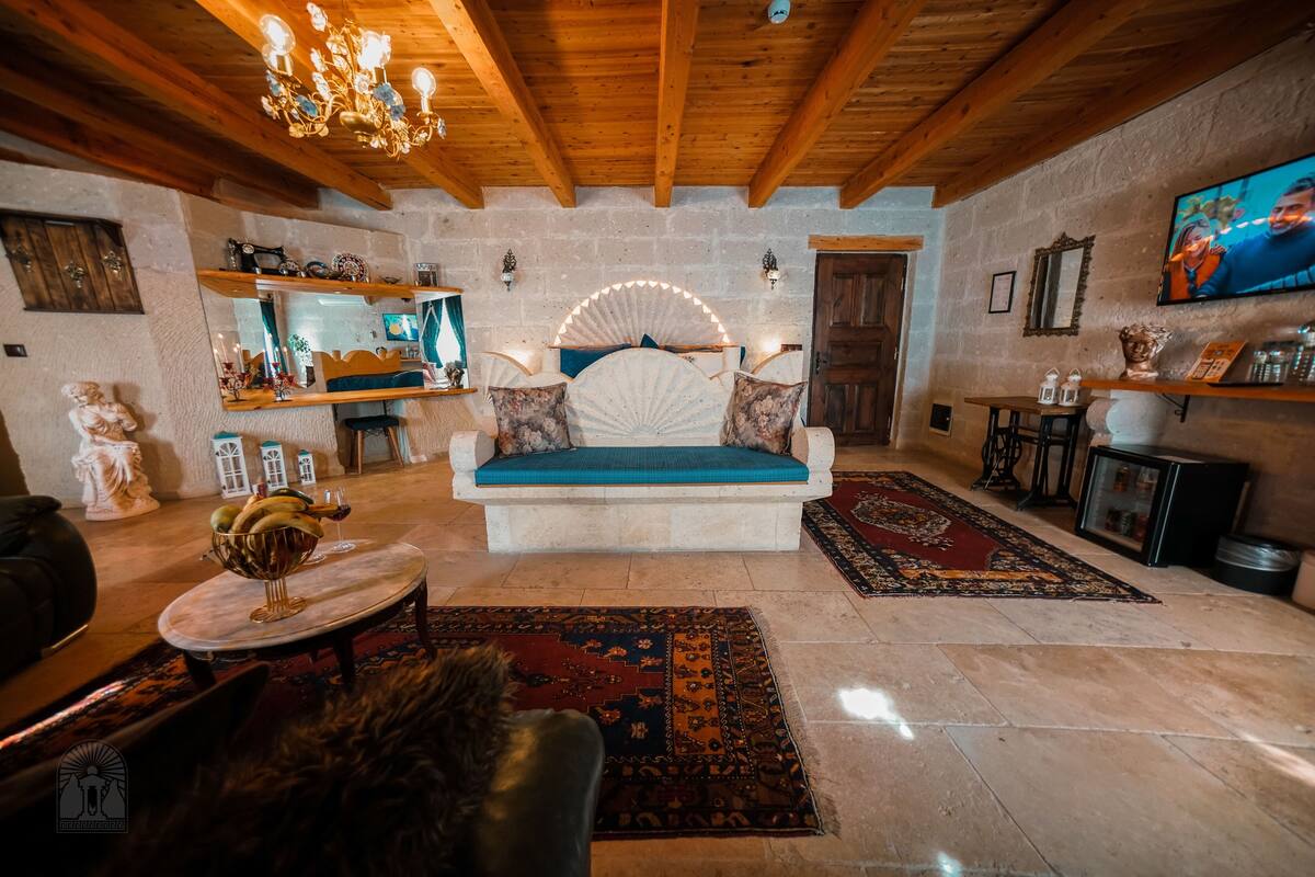 A cozy room with a stone wall, wooden ceiling beams, a large bed with a blue mattress, a chandelier, and a television on the wall.