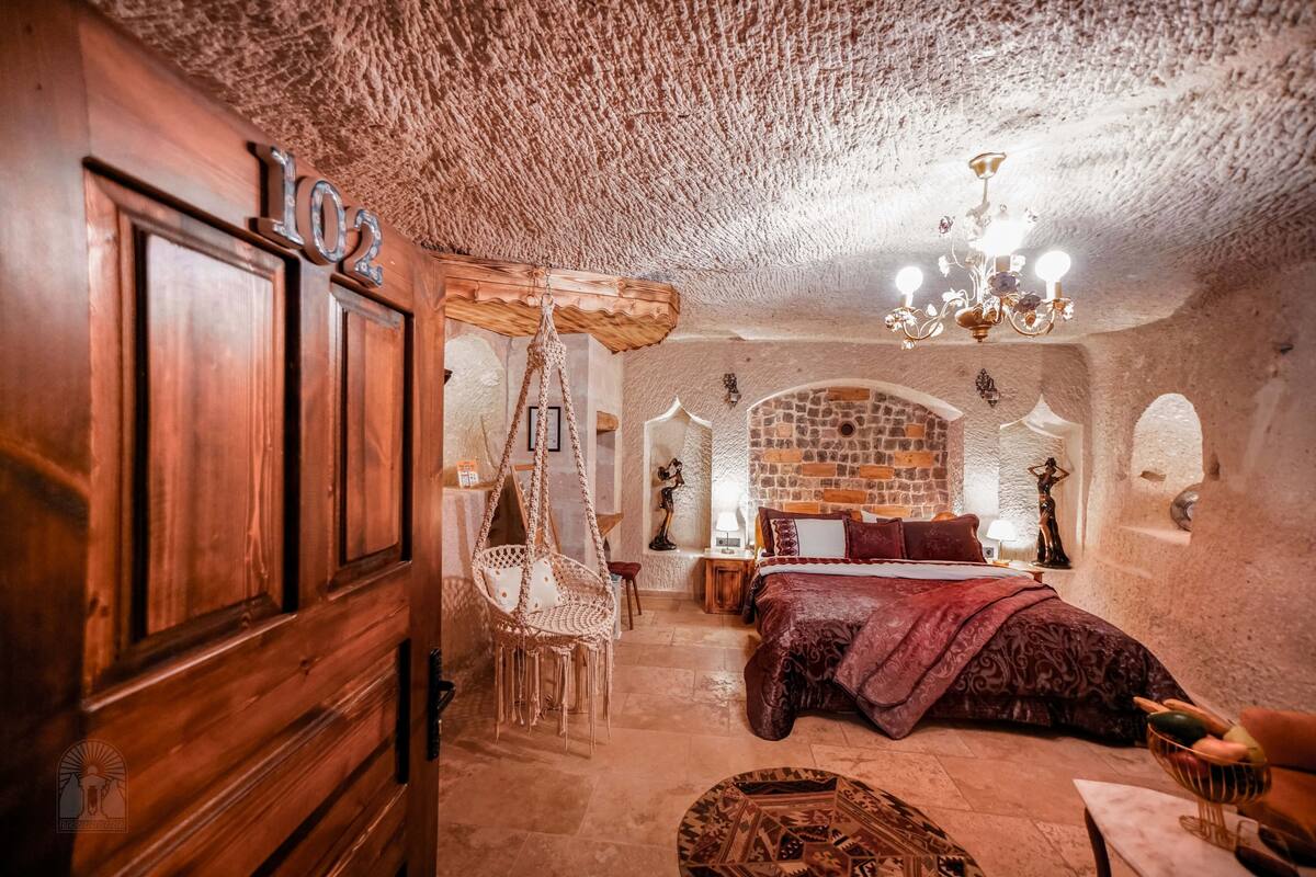 A rustic room with a wooden door labeled "102," a hanging chair, a bed with a red blanket, and a chandelier.
