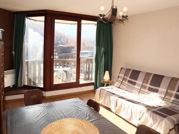 A room with a bed, a table, a chair, a lamp, and a view of a building through large windows.