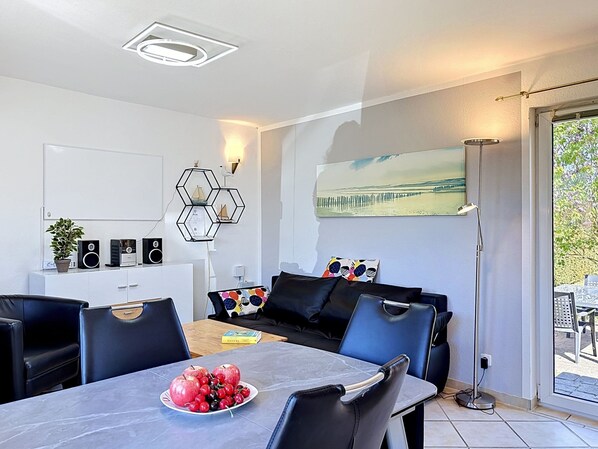 Een moderne woonkamer met een eethoek, een bank, een bijzettafeltje met fruit, en een schilderij aan de muur.