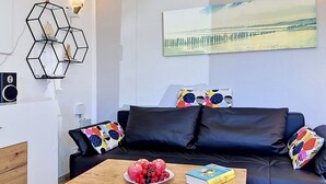 Una sala de estar moderna con un sofá de cuero negro, una mesa de centro de madera con fruta y un libro, una decoración geométrica en la pared y una pintura de un muelle.
