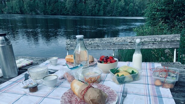A picnic setup on a wooden dock by a lake with a tablecloth, food, and drinks. - 2-bed Apartment Overlooking the River (Avesta)