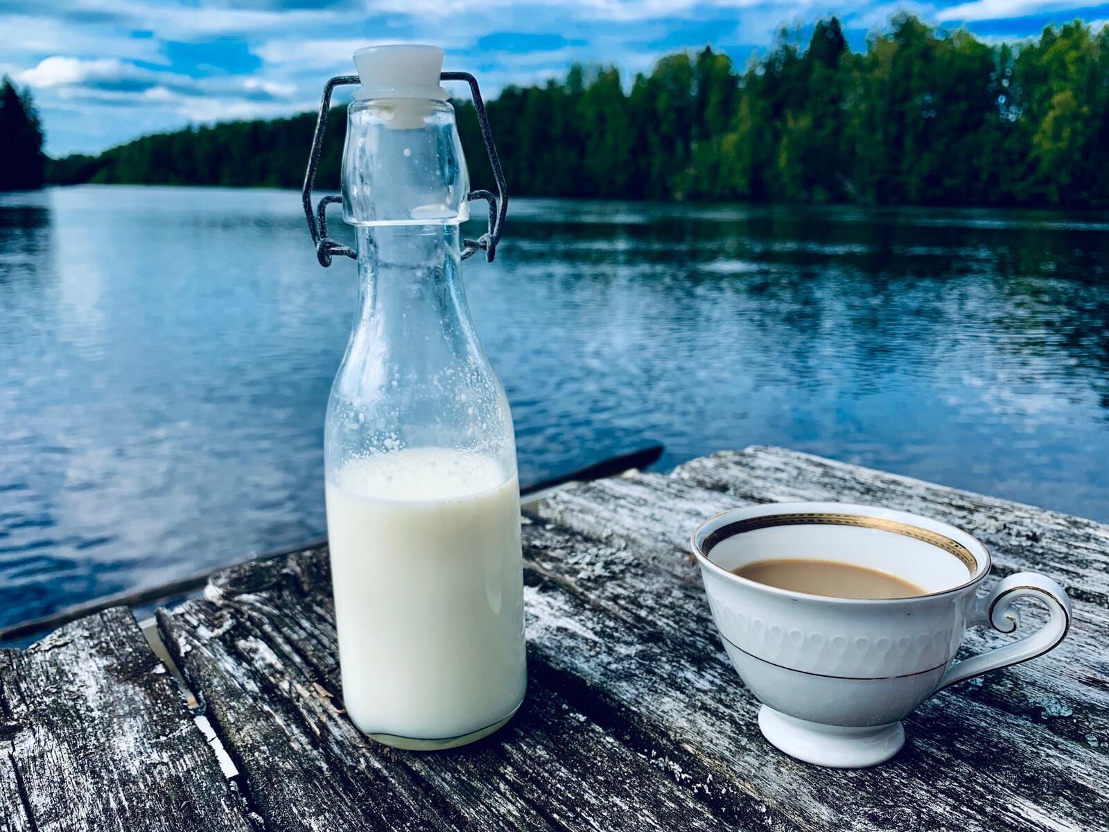 A glass milk bottle and a white cup on a wooden surface by a lake.
