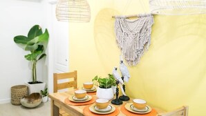 Un coin repas avec une table en bois dressée pour quatre personnes, entourée de chaises. On y trouve des décorations murales en macramé et une chaise en macramé. Un ara bleu et jaune est perché sur un support.