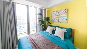 Une chambre avec un grand lit, une fenêtre avec des rideaux, un mur jaune et un tableau abstrait aux couleurs vives.