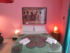 Quarto com cama, travesseiros, um quadro na parede, mesa de cabeceira com abajur e uma toalha dobrada na cama.