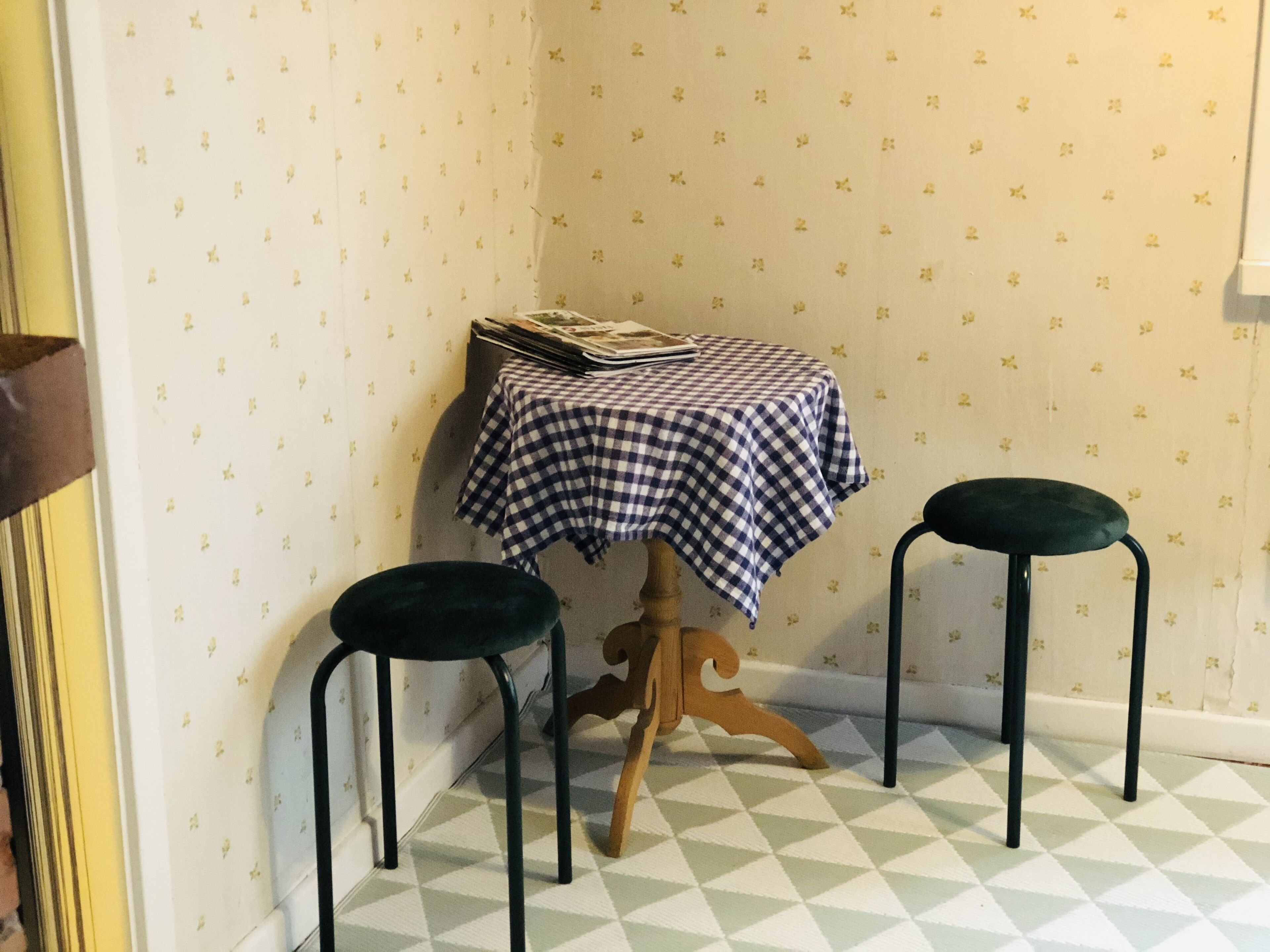A corner room with a checkered tablecloth, two stools, and patterned wallpaper.