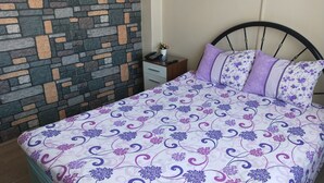 Un letto rifatto con cura, con una coperta a motivi floreali, una testiera in metallo nero e un piccolo comodino con una lampada e un vaso di fiori.