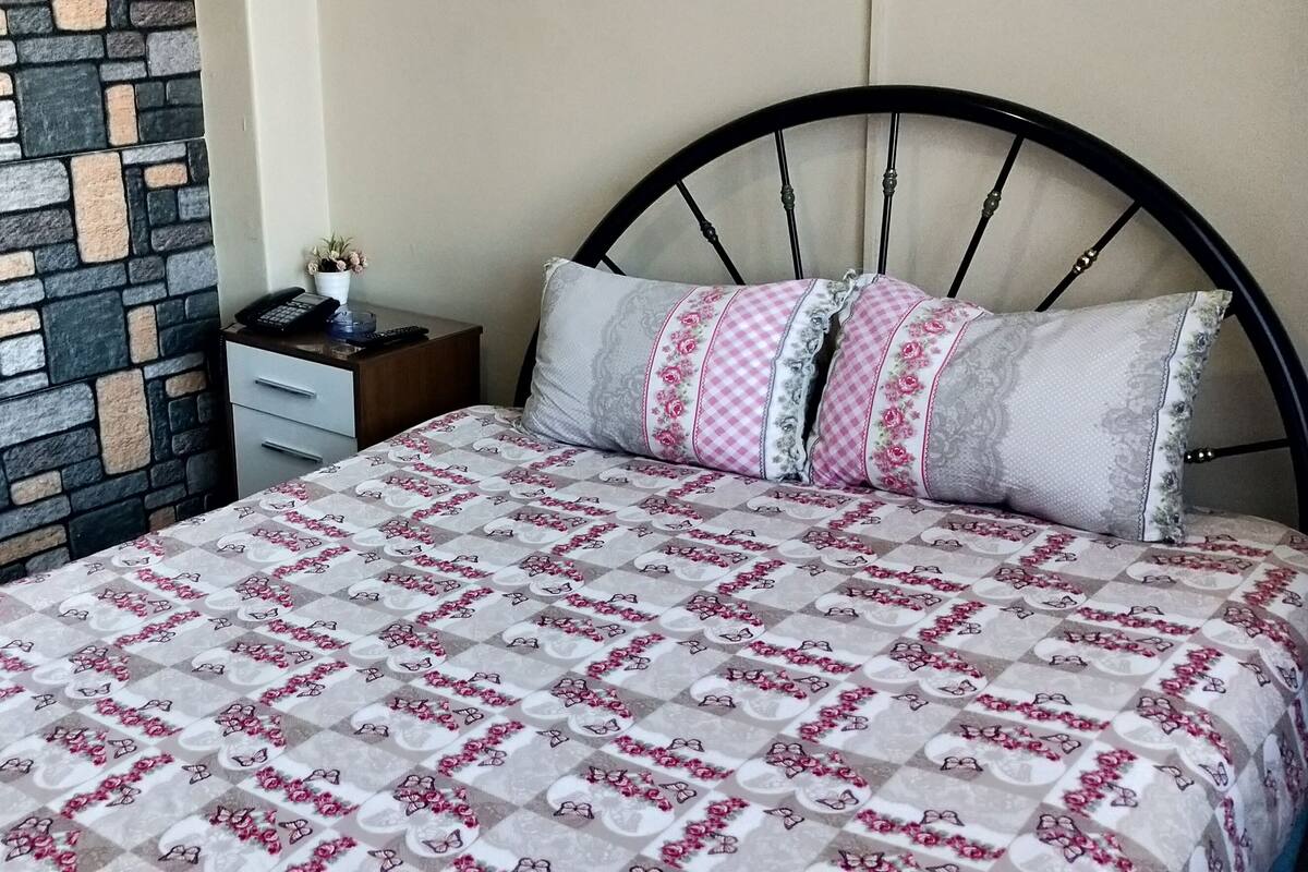 Un dormitorio con pared de piedra estampada, cama con colcha floral y mesita de noche con teléfono y una planta pequeña.