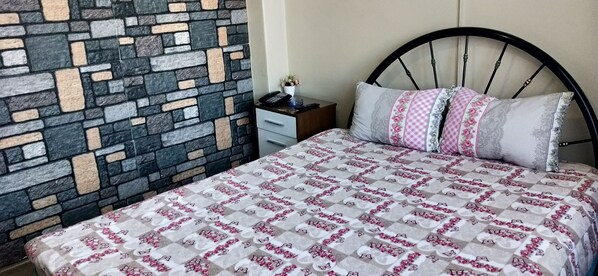 A bedroom with a stone-patterned wall, a bed with a floral bedspread, and a nightstand with a phone and a small plant. - Otel Cinar (Erzurum)