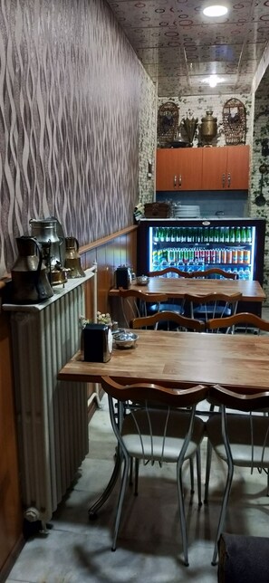 A dining area with a wooden table and metal chairs, a refrigerator stocked with beverages, and decorative items on the wall. - Otel Cinar (Erzurum)